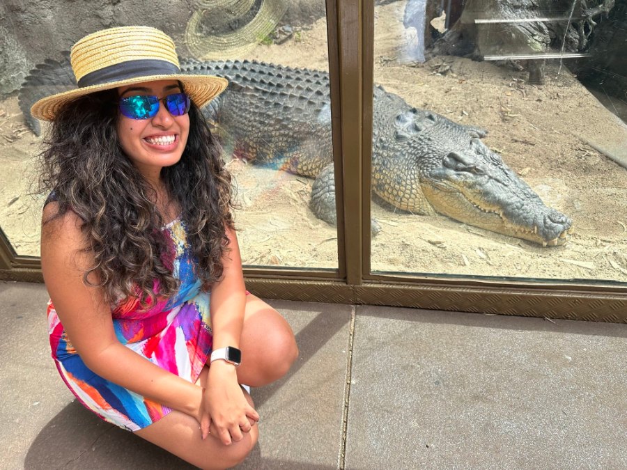 Meenu crouching by the glass, smiling, with a big estuarine crocodile behind her