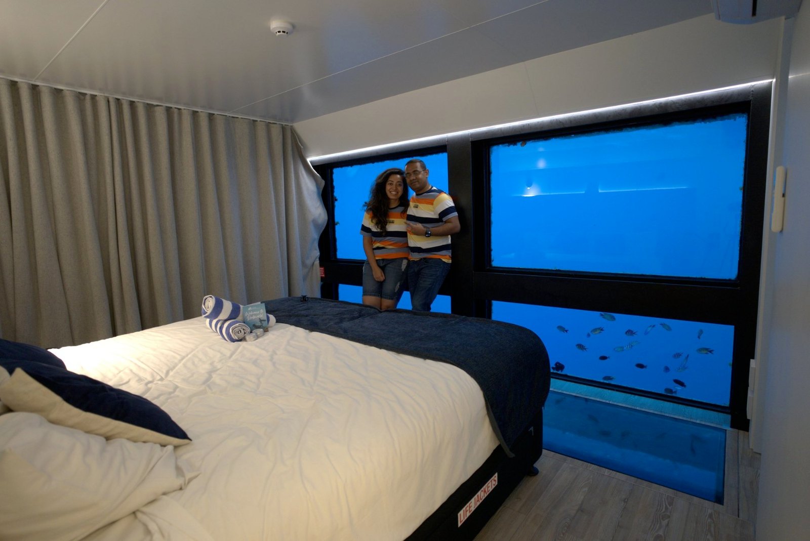 Meenu and Praddy at the window of our underwater room, with fish in the blue water outside