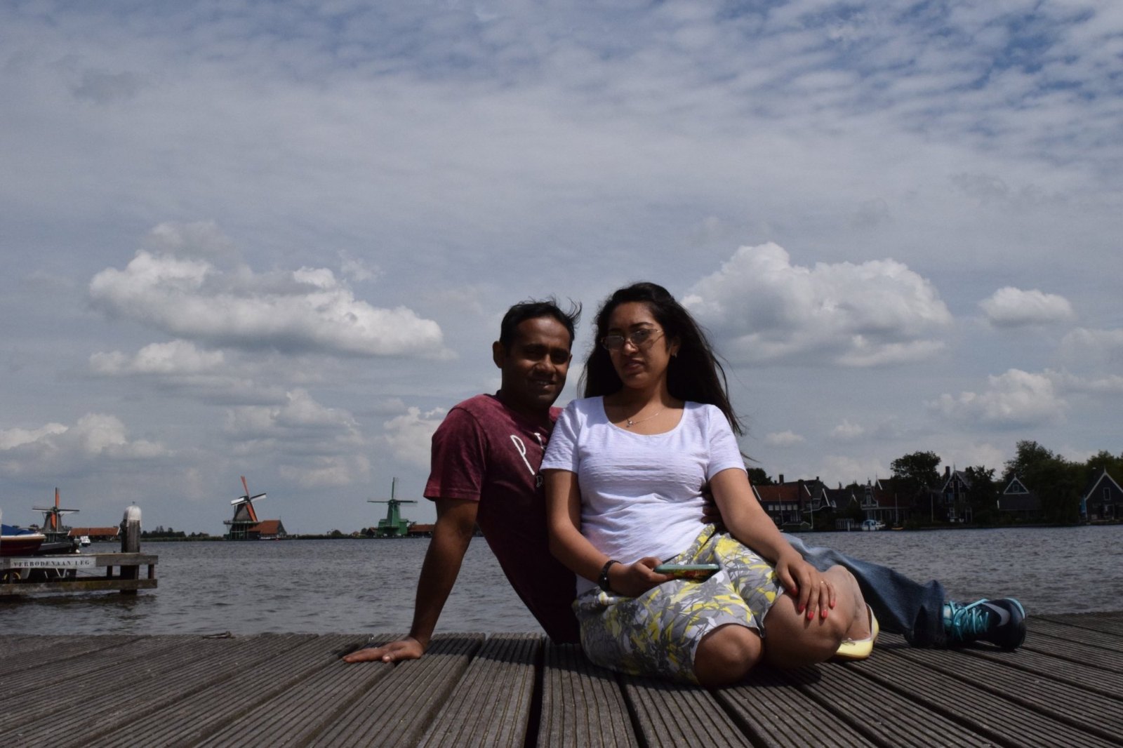Praddy and Meenu sitting on a wooden jetty on the Zaan, with windmills along the far bank