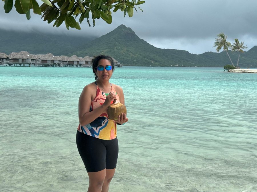 Meenu standing in the shallow lagoon with a fresh coconut, overwater villas behind