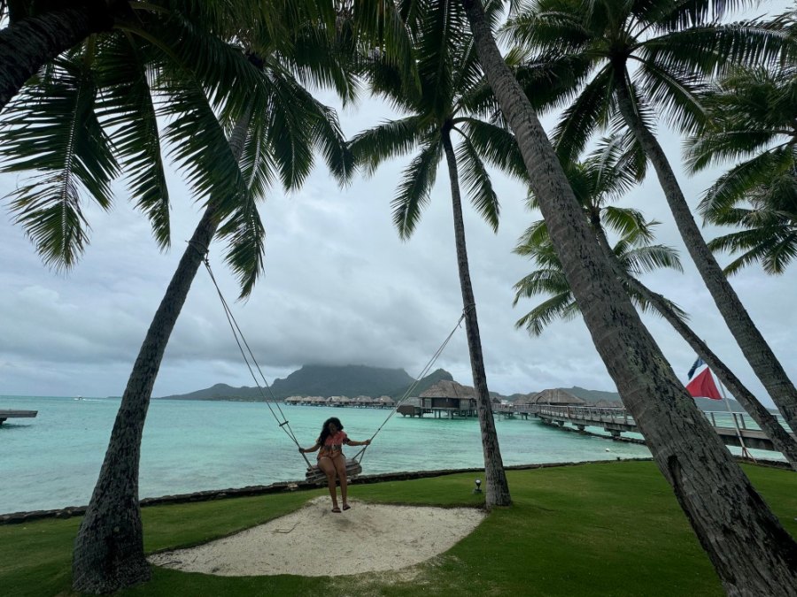 Meenu on a rope swing between leaning palms, with overwater villas and a cloud-capped Otemanu beyond