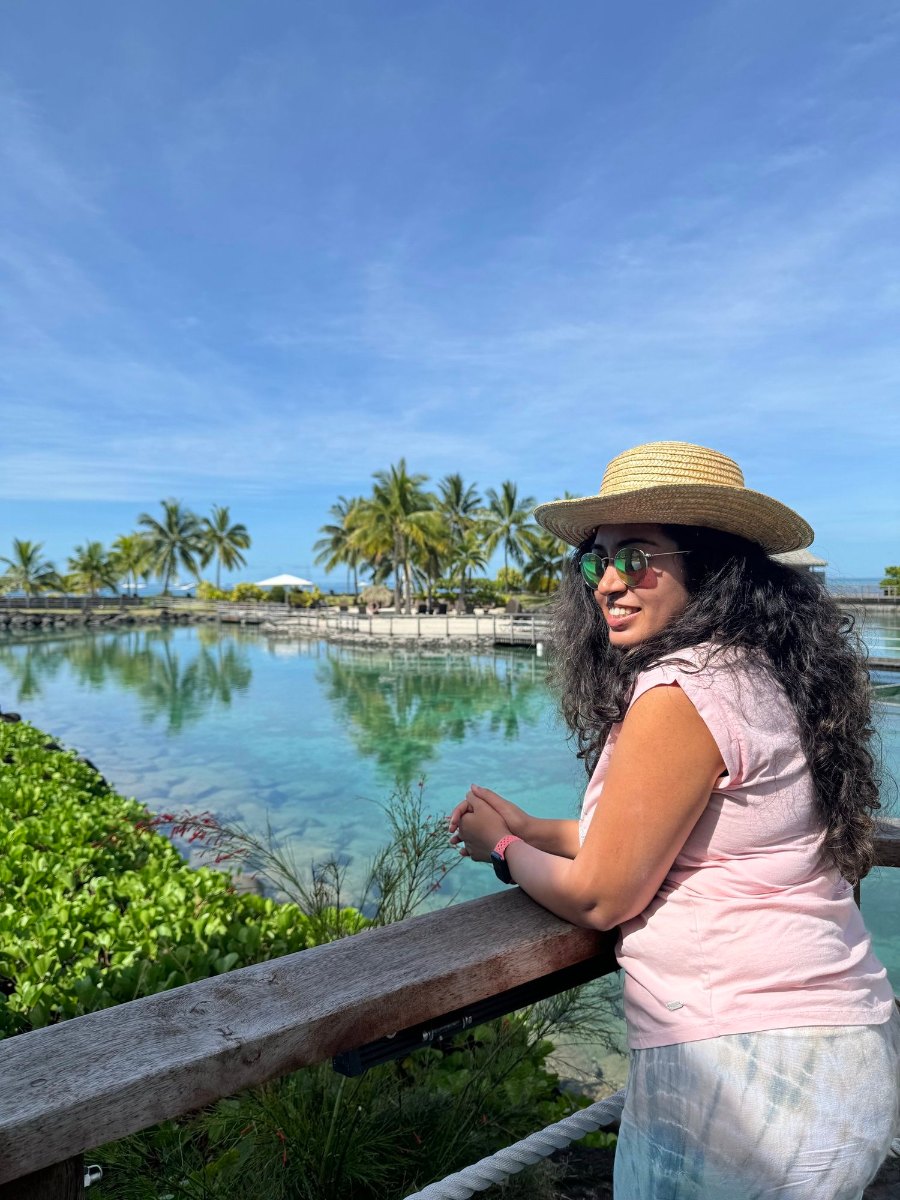 Meenu in a straw hat beside the hotel's lagoon, with palms reflected in the water
