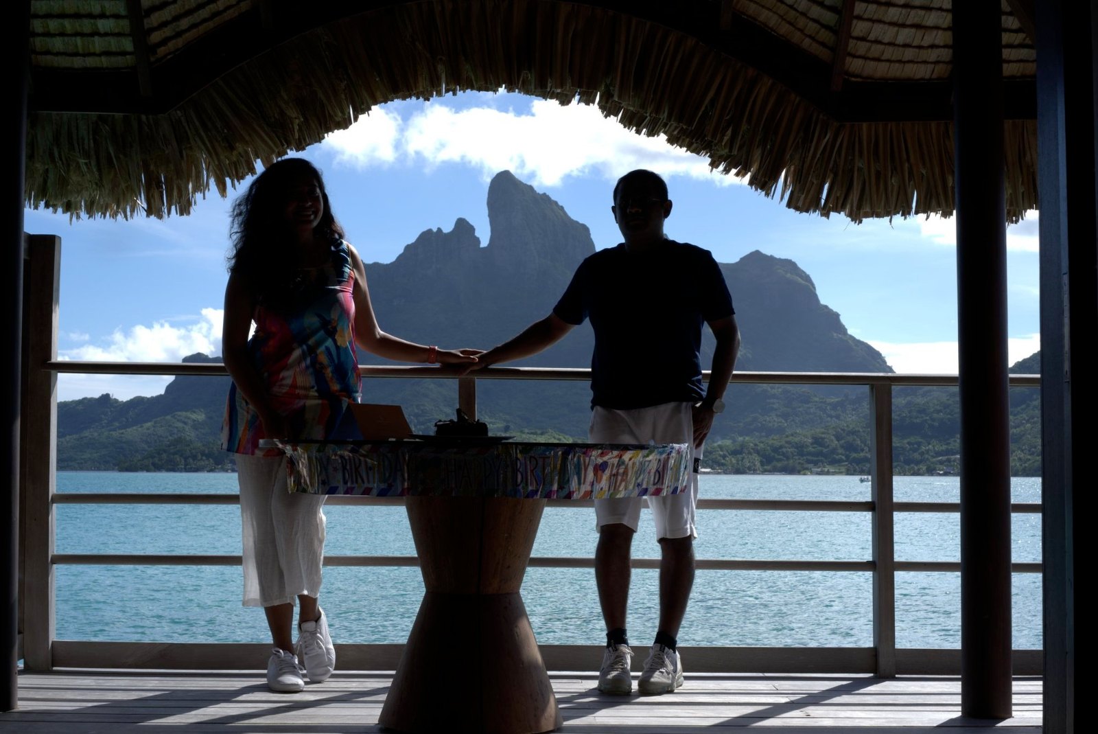 Meenu and Praddy in silhouette, holding hands at a rail under a thatched roof, with Otemanu across the lagoon
