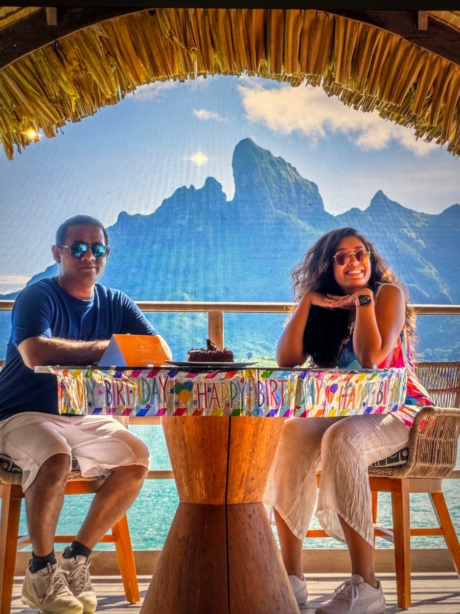 Meenu and Praddy behind a Happy Birthday banner and a chocolate cake, with Otemanu behind them