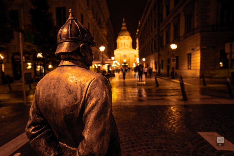 A bronze policeman in an old-fashioned helmet, seen from behind, looking up a lamplit street to the Basilica