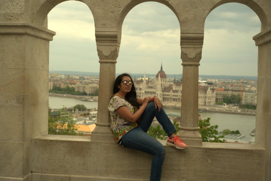 Meenu sitting in a stone arch at Fisherman's Bastion, with the Parliament framed across the river