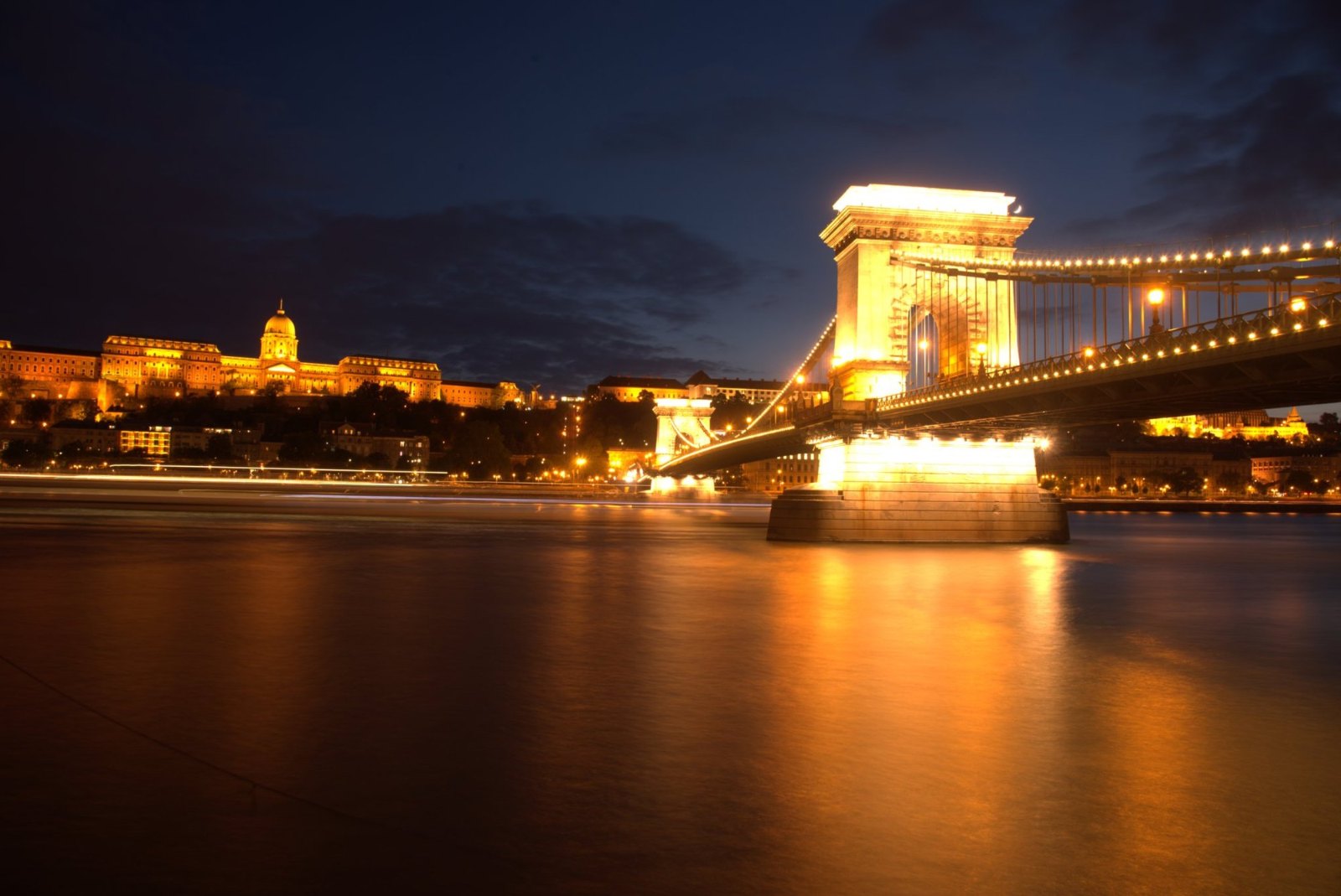 A tower of the Chain Bridge lit gold at night, with Buda Castle glowing on the hill and the lights reflected in the river