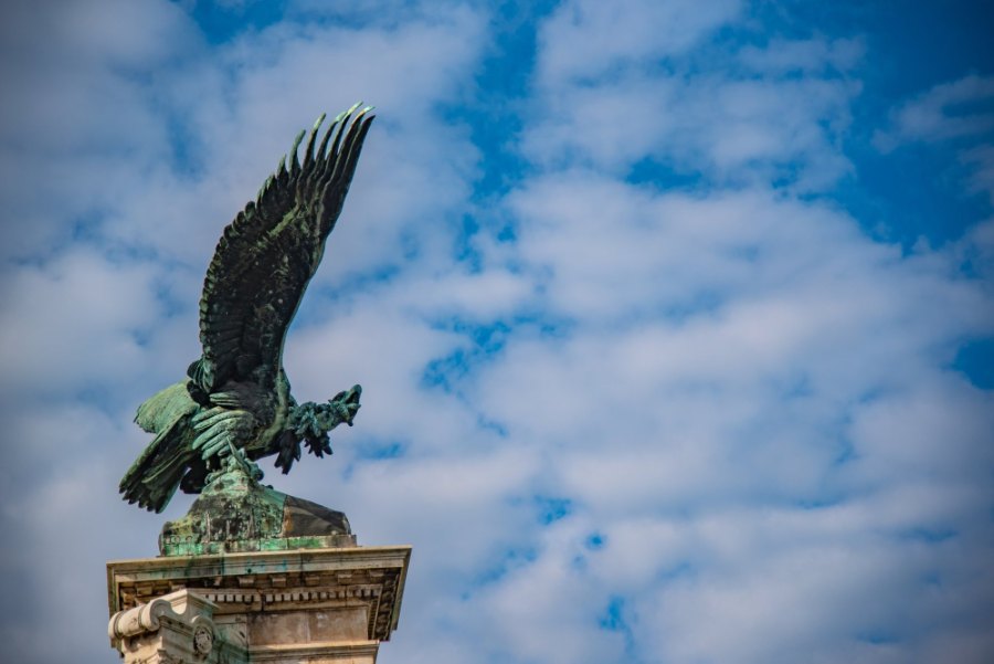 The bronze Turul bird spreading its wings on a stone pedestal, against a blue sky with clouds