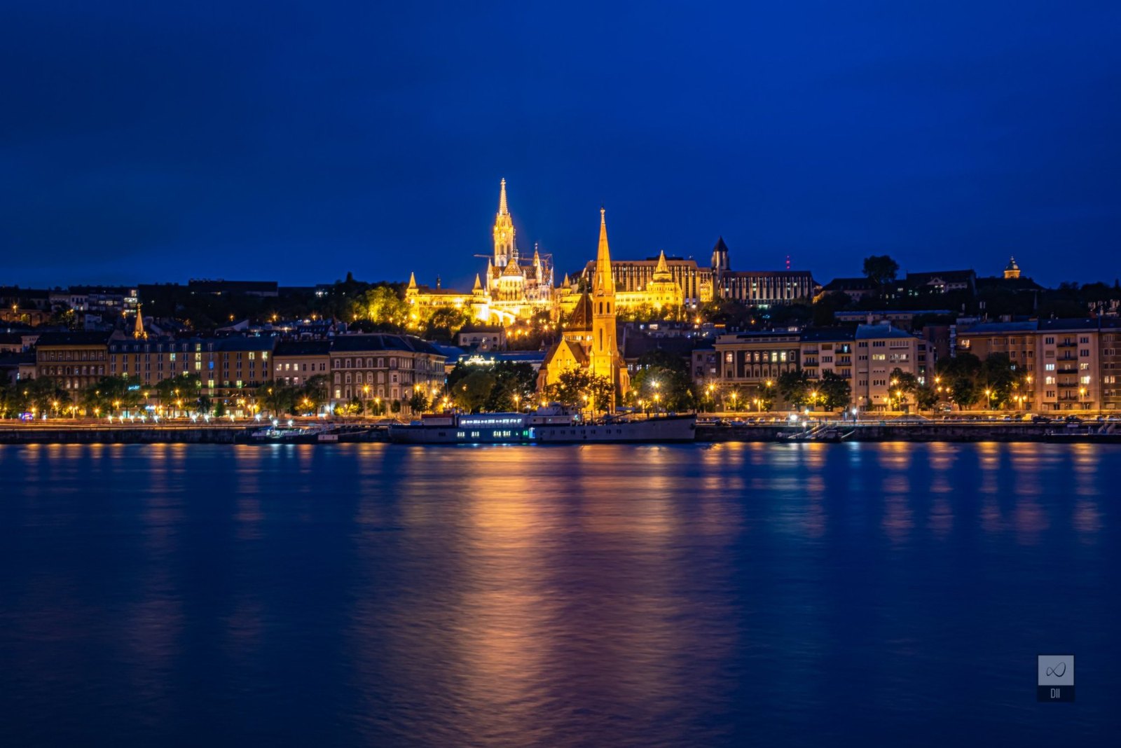 Matthias Church and Fisherman's Bastion lit gold above the Buda riverfront, across the Danube at blue hour