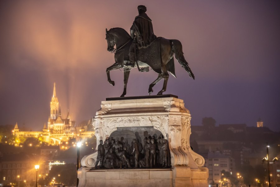 A bronze rider on a carved stone plinth, with Fisherman's Bastion glowing across the river and beams of light in the mist