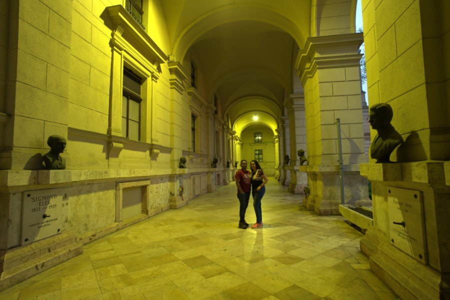 Meenu and Praddy standing in a long arcade lit yellow, with bronze busts along the walls