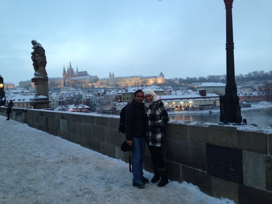 The two of us on snowy Charles Bridge at dusk beside a statue, with Prague Castle lit across the river