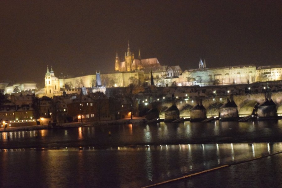 Prague Castle lit up at night above the arches and lamps of Charles Bridge, reflected in the river