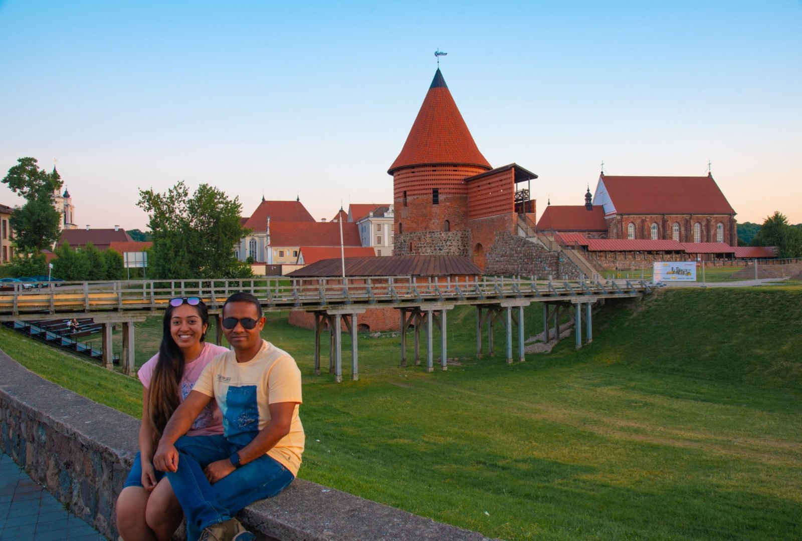 Meenu and Praddy on a low wall in front of Kaunas Castle's round red-brick tower at sunset