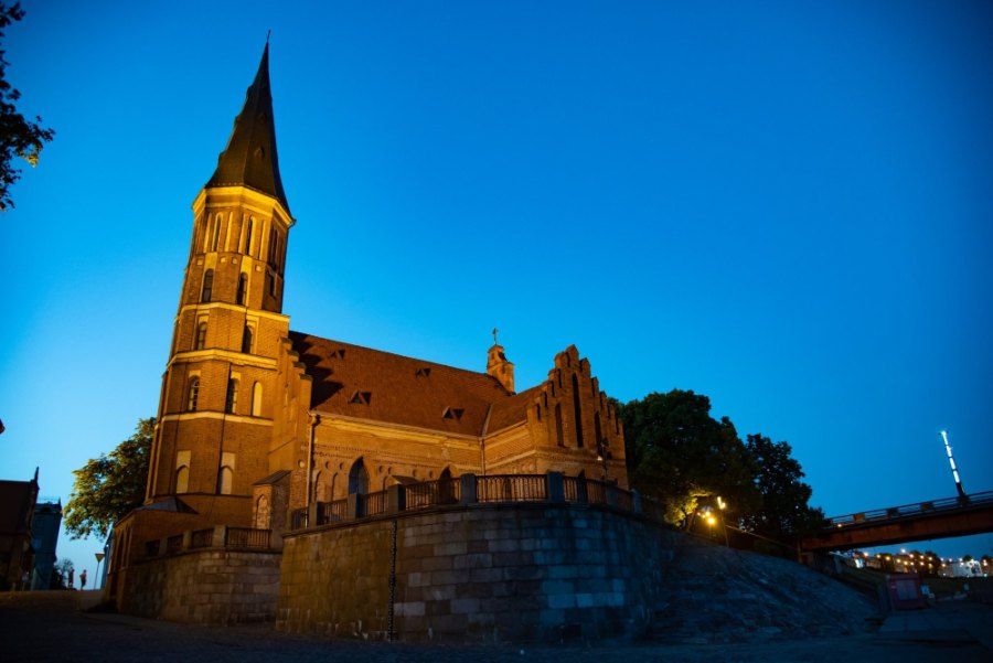 The red-brick Church of Vytautas the Great lit up on the riverbank at night, beside the red bridge