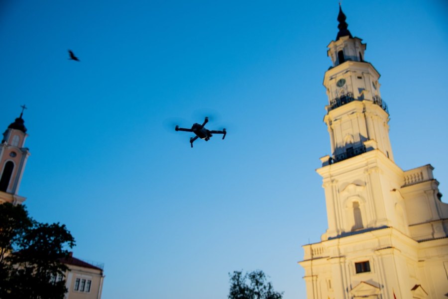 A drone in flight beside the floodlit white tower of Kaunas Town Hall
