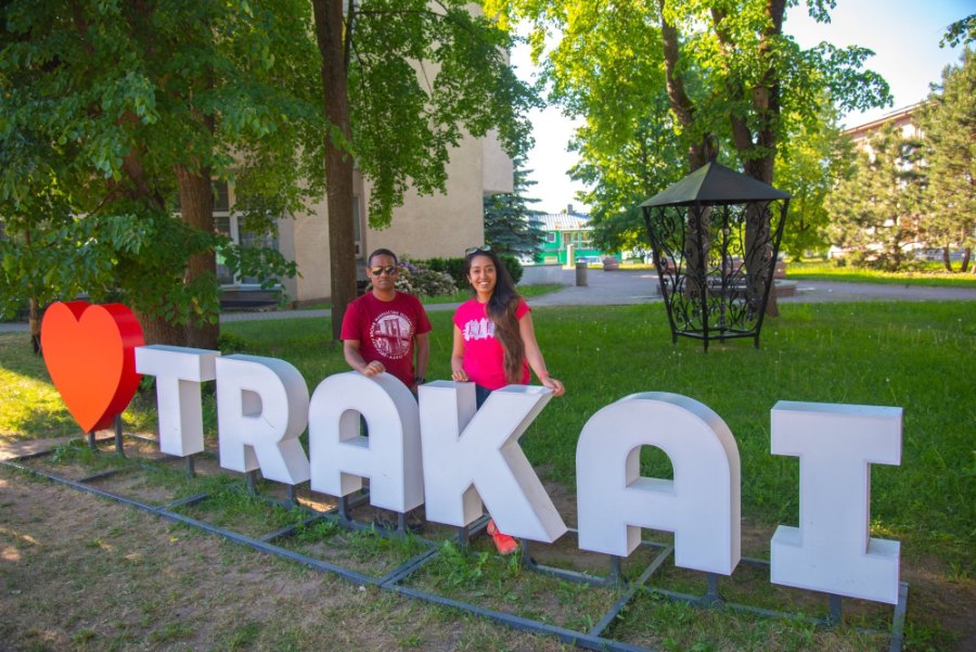 Meenu and Praddy behind big white letters spelling TRAKAI, with a red heart, on the grass under the trees