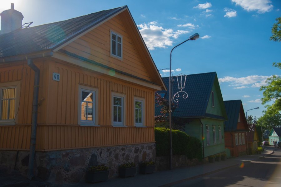 A yellow wooden house with three windows facing the street, and more wooden houses beyond, one painted green