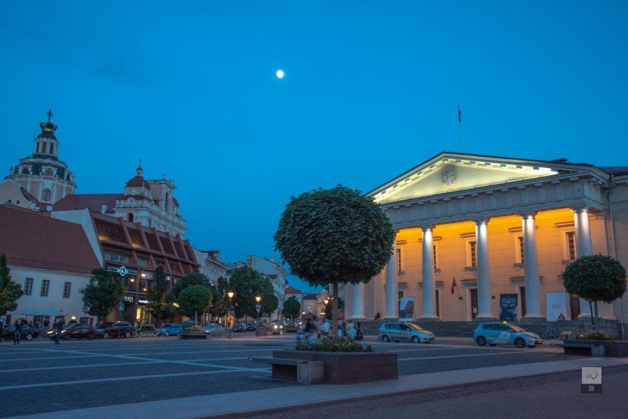 The Town Hall's columns lit gold on Town Hall Square at dusk, with the moon above and St Casimir's dome to the left