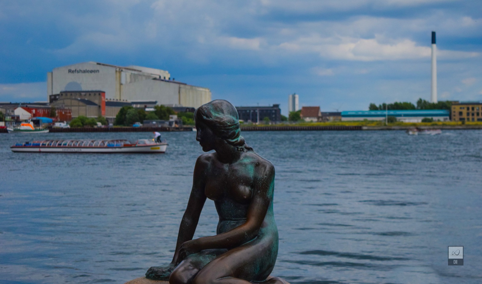 The Little Mermaid on her rock, looking out over the harbour as a tour boat goes by