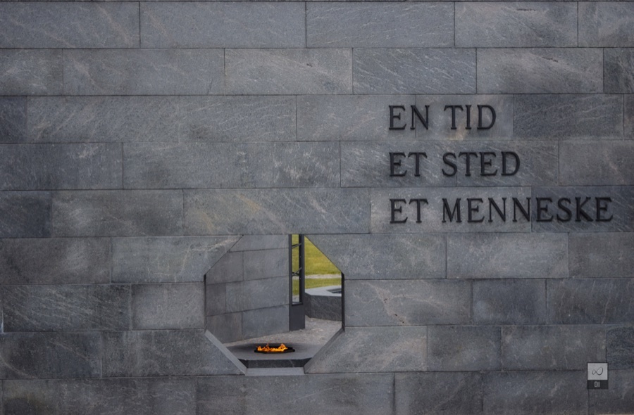 A grey stone memorial wall carved with the words En tid, et sted, et menneske, above a flame burning in a niche