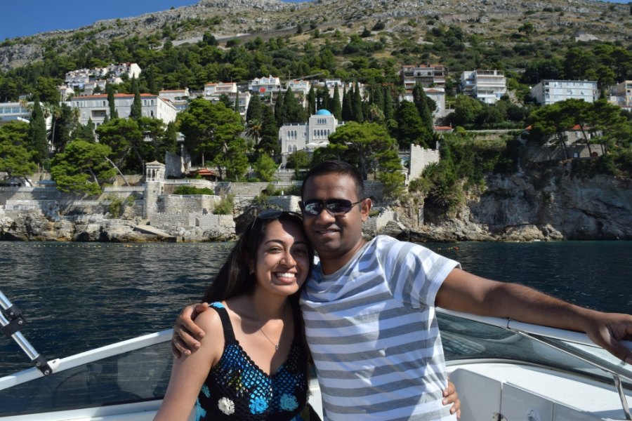 Meenu and Praddy arm in arm on the speedboat, with villas and pines along the shore behind