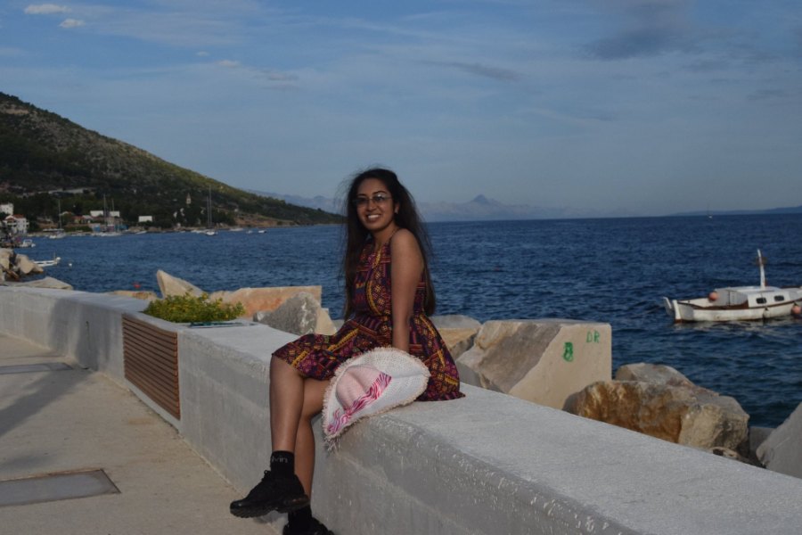 Meenu sitting on the white sea wall of the promenade, with the sea and a small boat behind