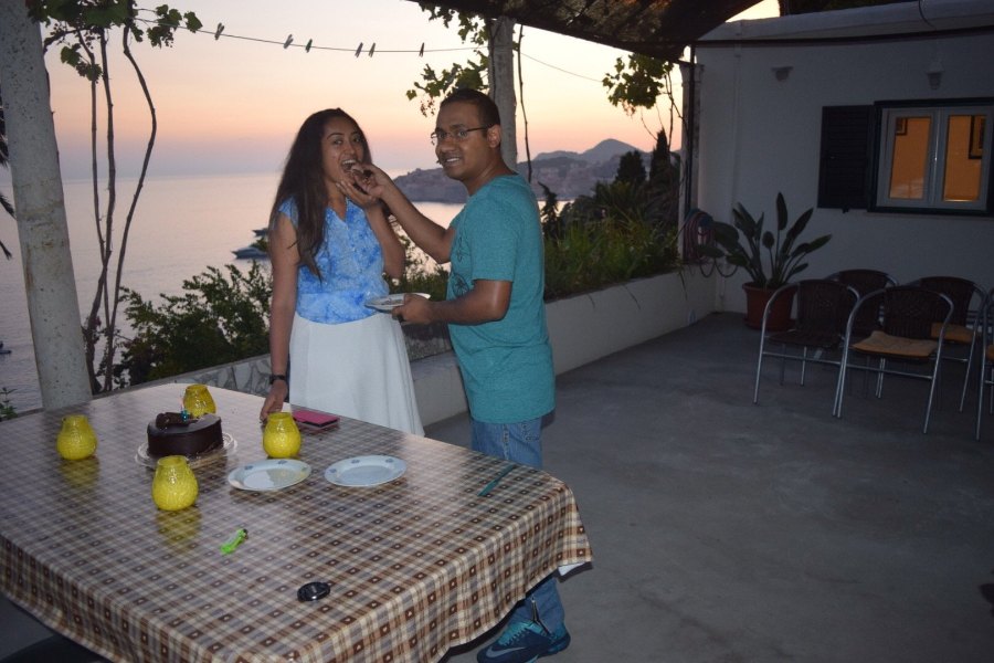 Praddy feeding Meenu a piece of birthday cake on the terrace at sunset, with the coast behind