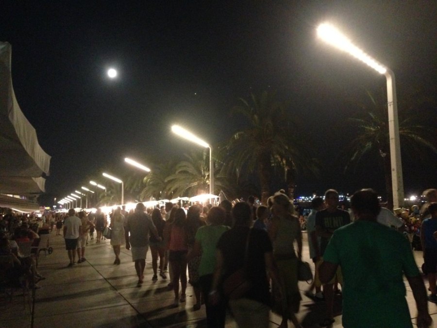 Crowds on the Riva at night under a full moon, between palms and tall street lamps