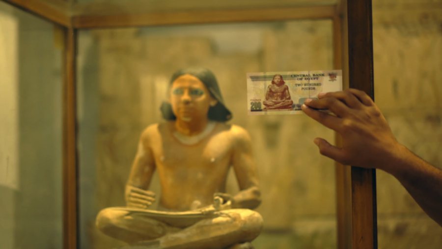 A 200-pound note held up beside the seated scribe statue printed on it