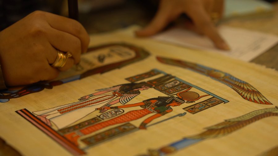 A hand with a gold ring tracing a painted scene on papyrus