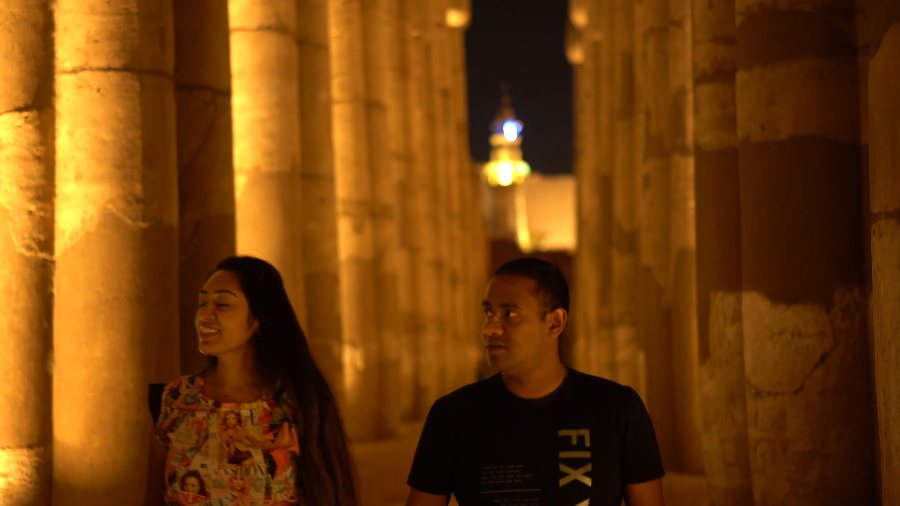 Meenu and Praddy walking between Luxor Temple's floodlit columns at night, with a lit minaret beyond