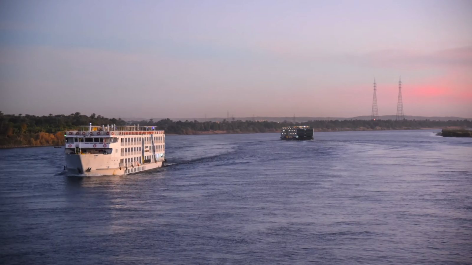 A white cruise boat sailing along the Nile at dusk, under a pink sky