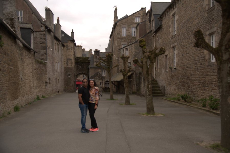 Meenu and Praddy in a wide stone street lined with pollarded trees, with an arched gateway at the end