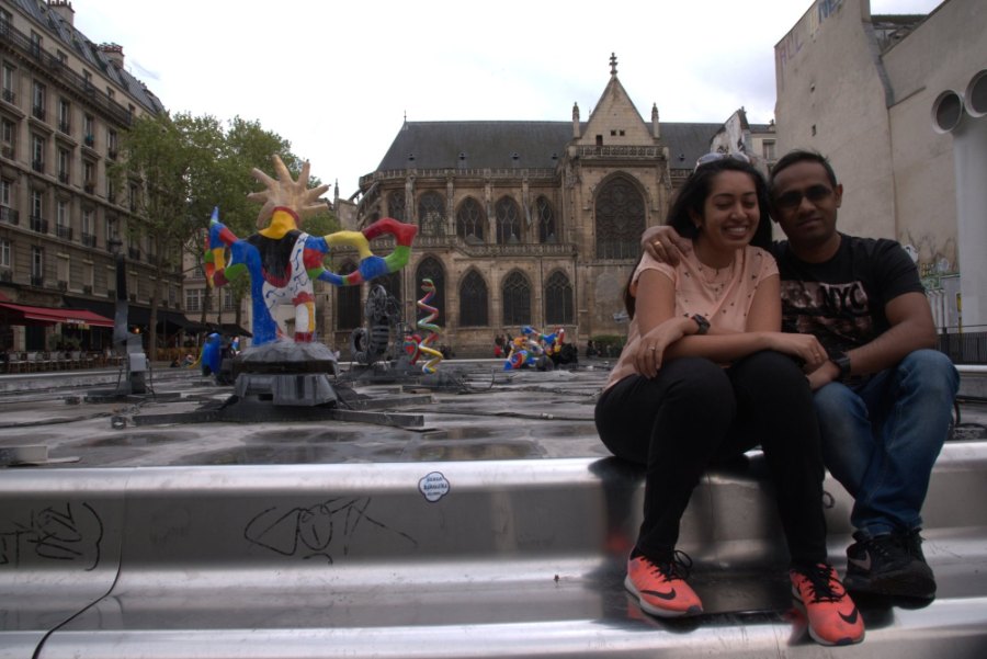 Meenu and Praddy sitting by the Stravinsky Fountain, with its bright sculptures and a Gothic church behind