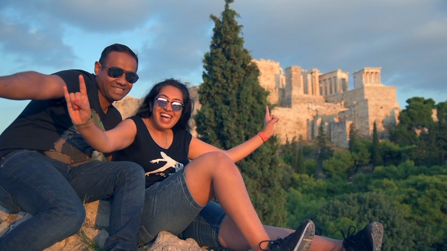 Meenu and Praddy laughing in a selfie on a rocky hill, with the Acropolis behind them
