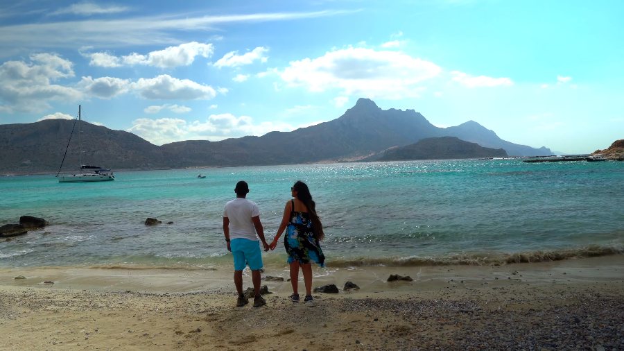 Meenu and Praddy walking hand in hand to the water's edge, with mountains across the lagoon