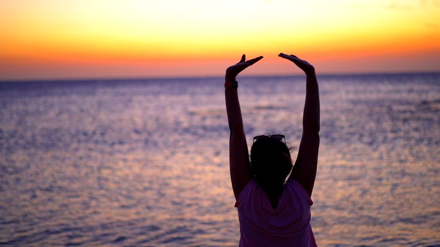 Meenu silhouetted against an orange sunset over the sea, arms raised above her head