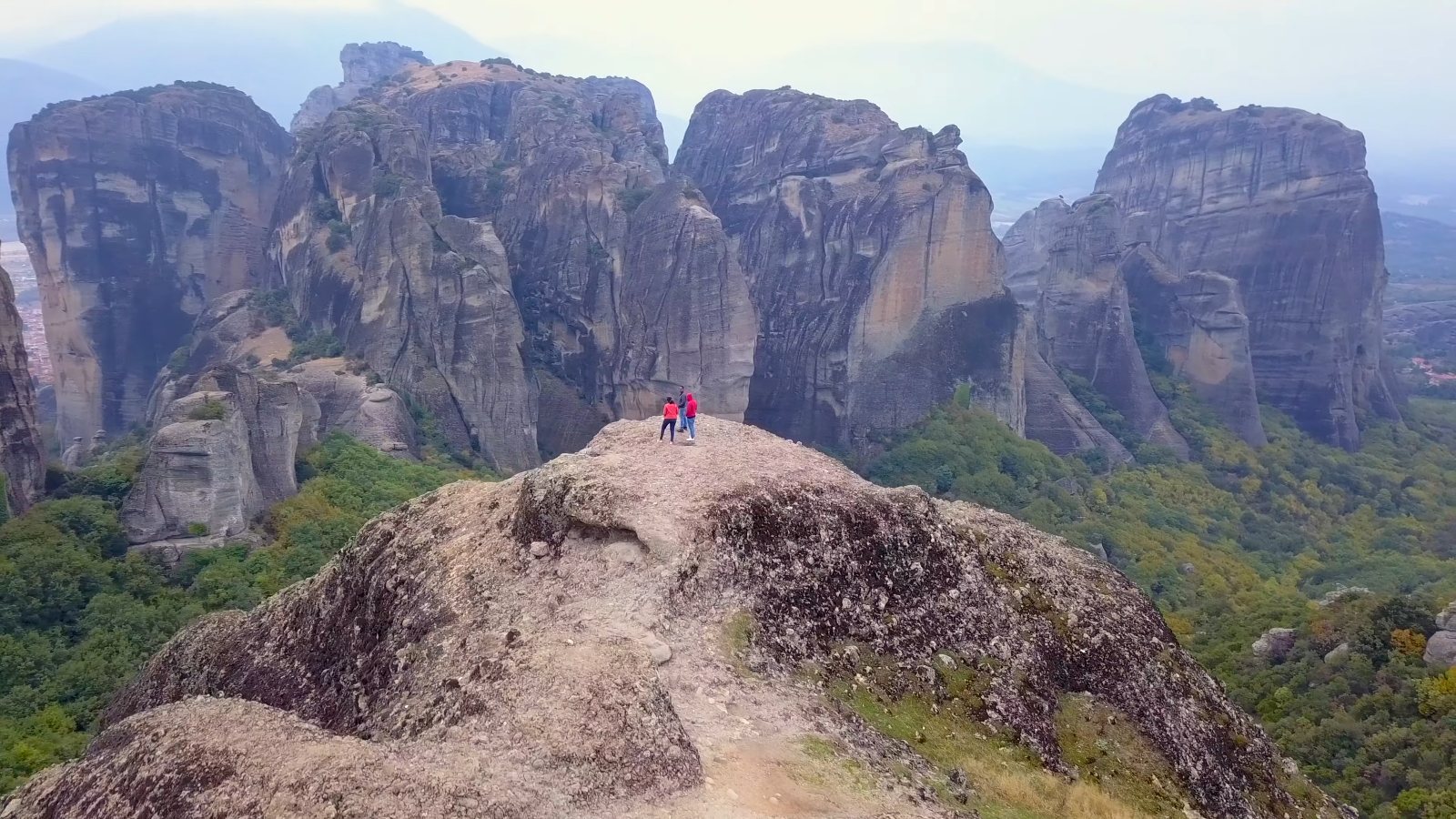 Drone view of Meenu and Praddy on a bare rock summit facing Meteora's rock towers
