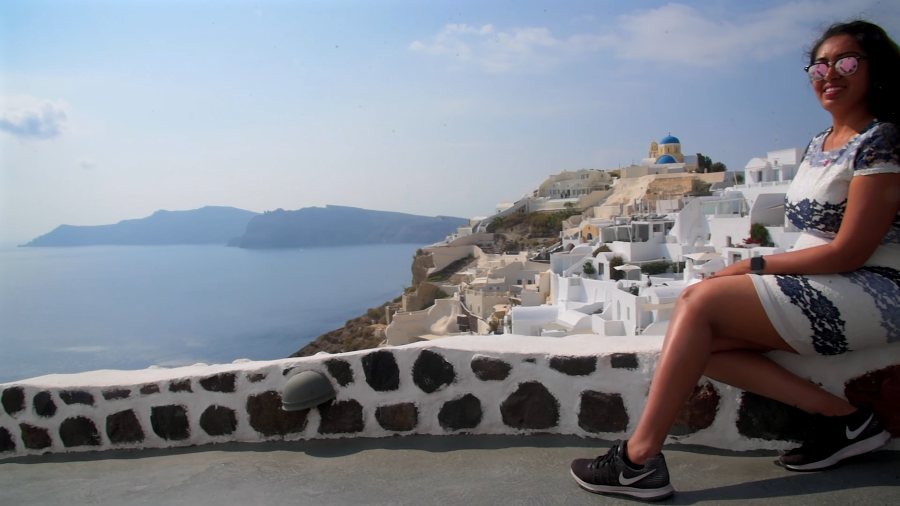 Meenu sitting on a whitewashed wall above Oia, with the caldera behind