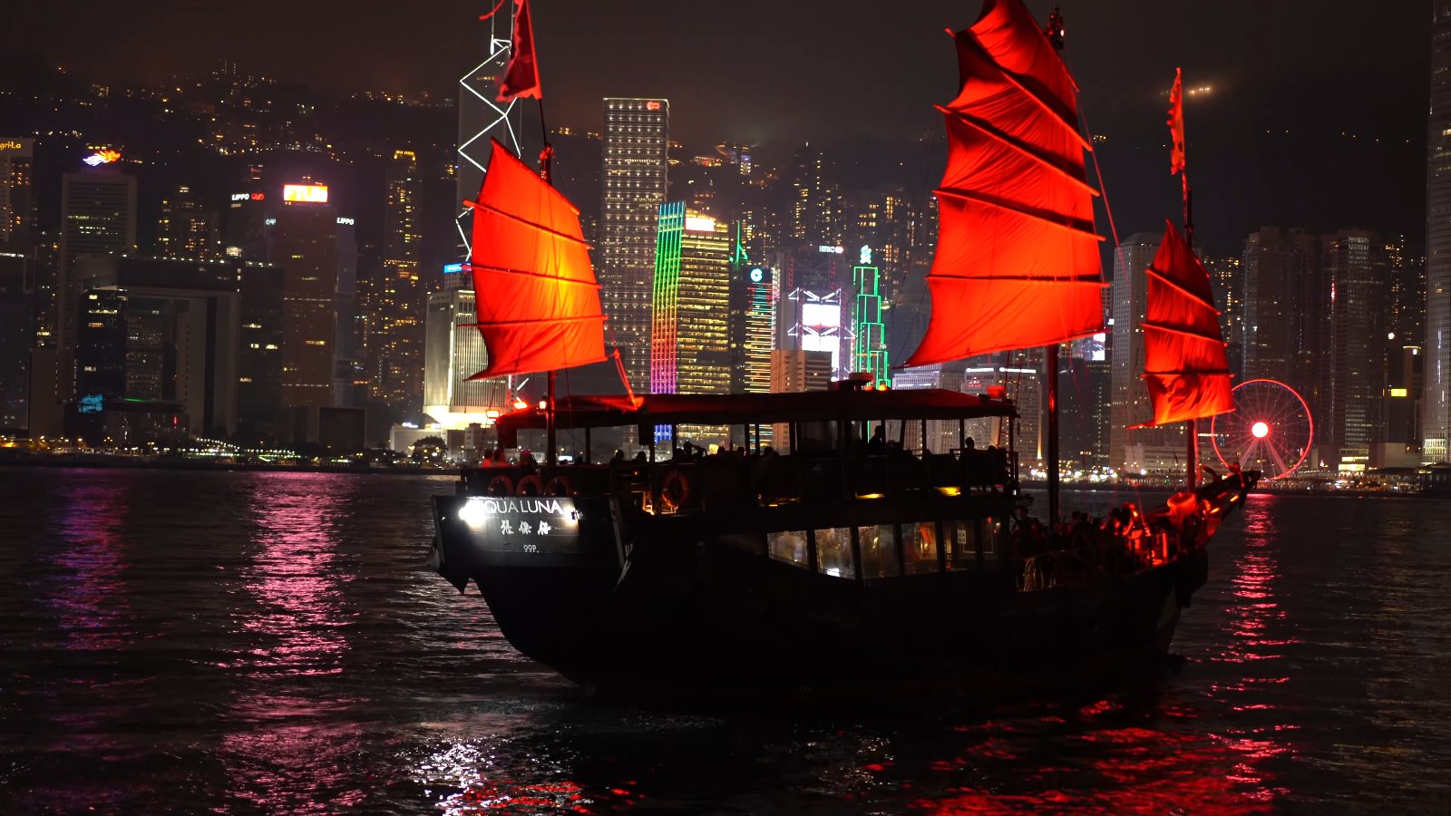 The Aqua Luna under red sails, passing the lit-up Bank of China Tower at night