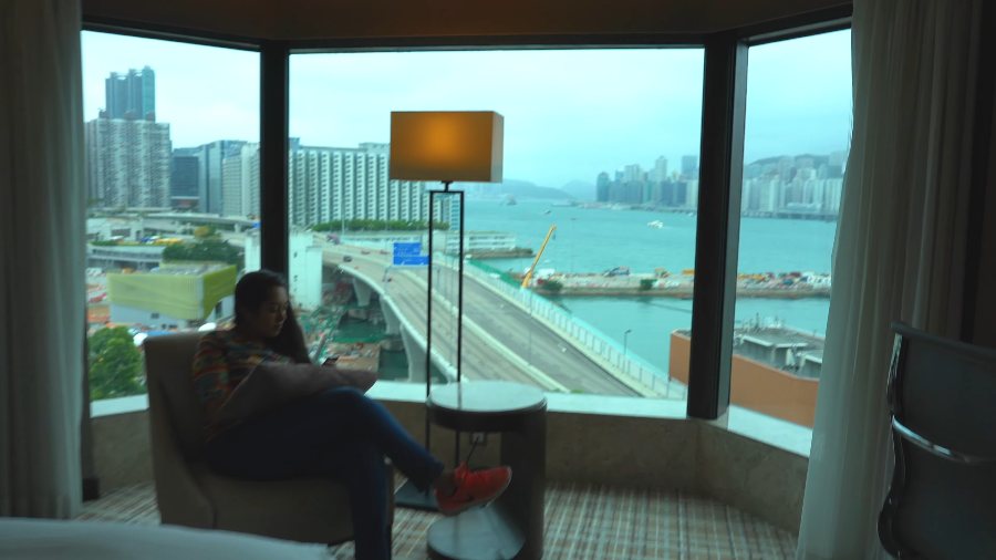 Meenu in an armchair at the corner window of our room, above a curving road and the harbour