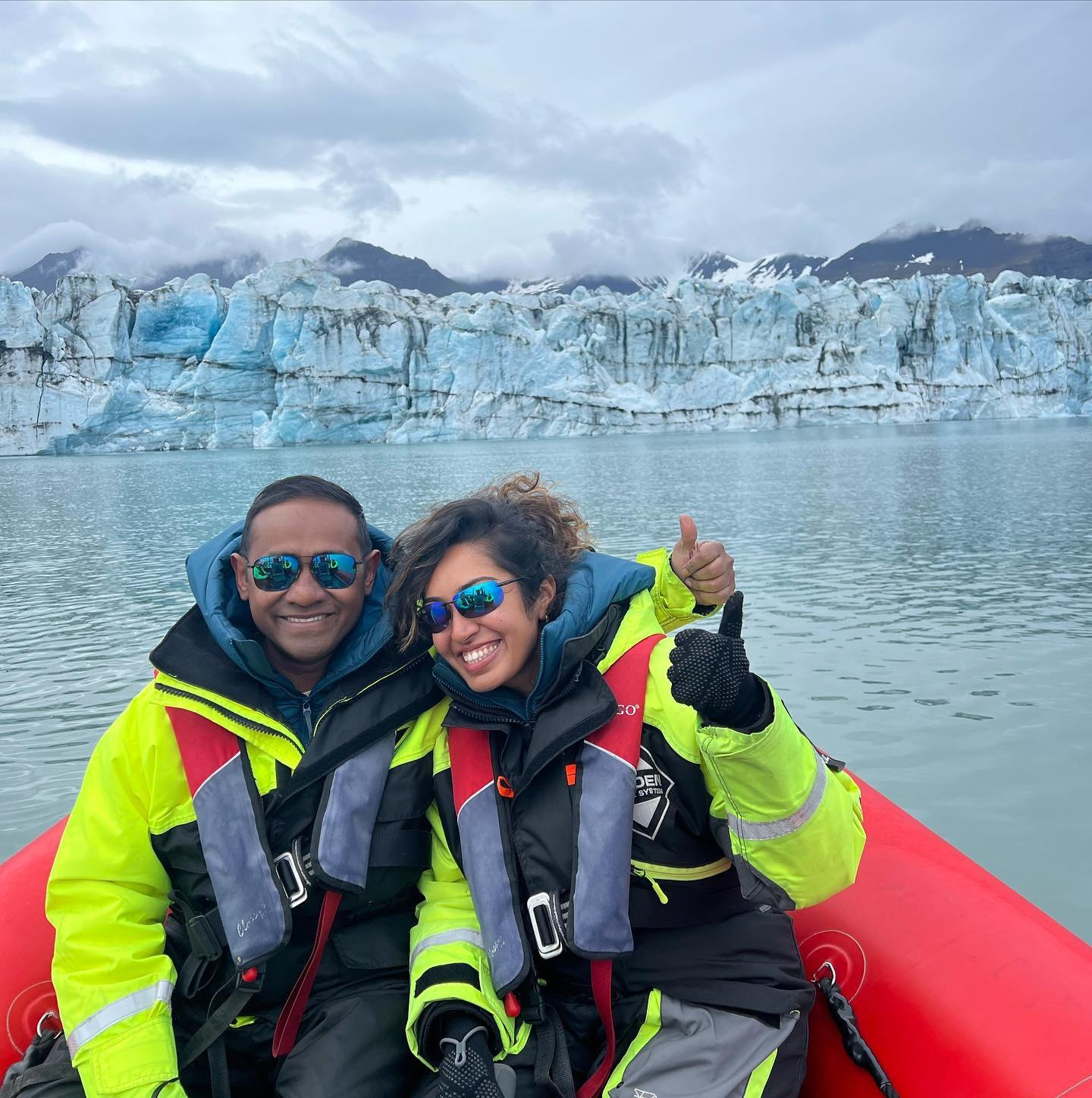 Meenu and Praddy in yellow flotation suits on a red zodiac on Jökulsárlón, with the glacier's blue-white face behind them