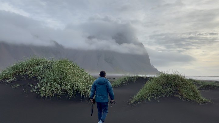 Praddy walking across black sand between grassy dunes at Stokksnes, toward a mountain wrapped in cloud