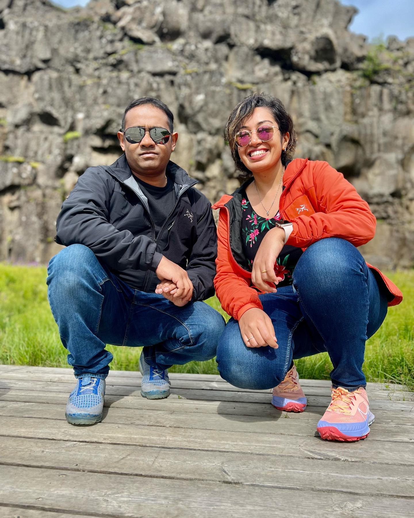 Meenu and Praddy crouching on a boardwalk at Þingvellir, in front of the rift's dark rock wall