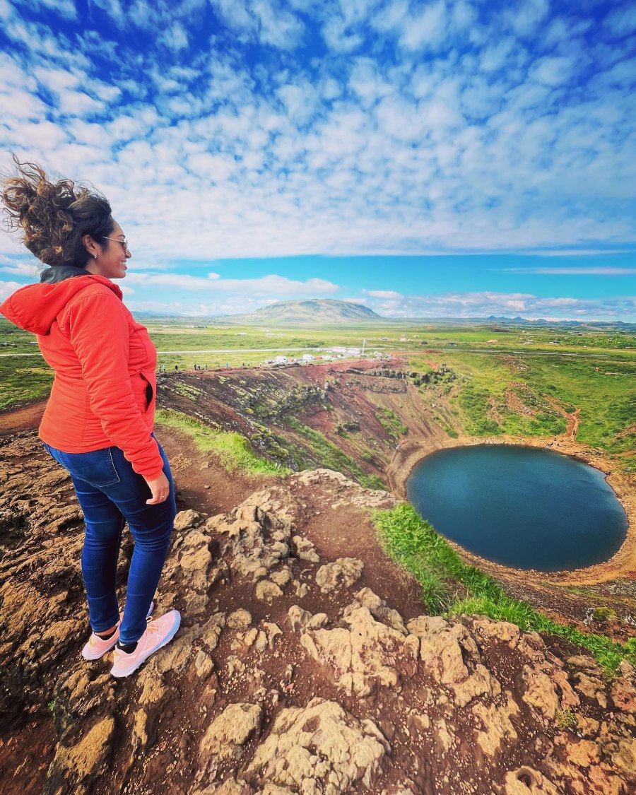Meenu in a red jacket on the rim of Kerið crater, her hair blowing, above the crater's blue lake