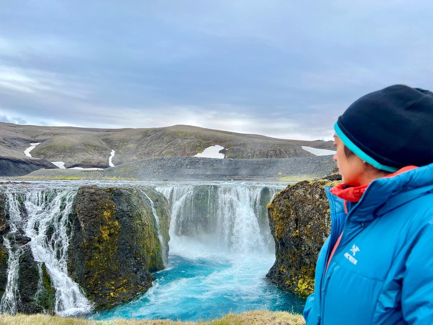 Meenu in a blue jacket and black hat beside Sigöldufoss, its streams pouring over mossy rock into a turquoise pool, with snow on the hills behind