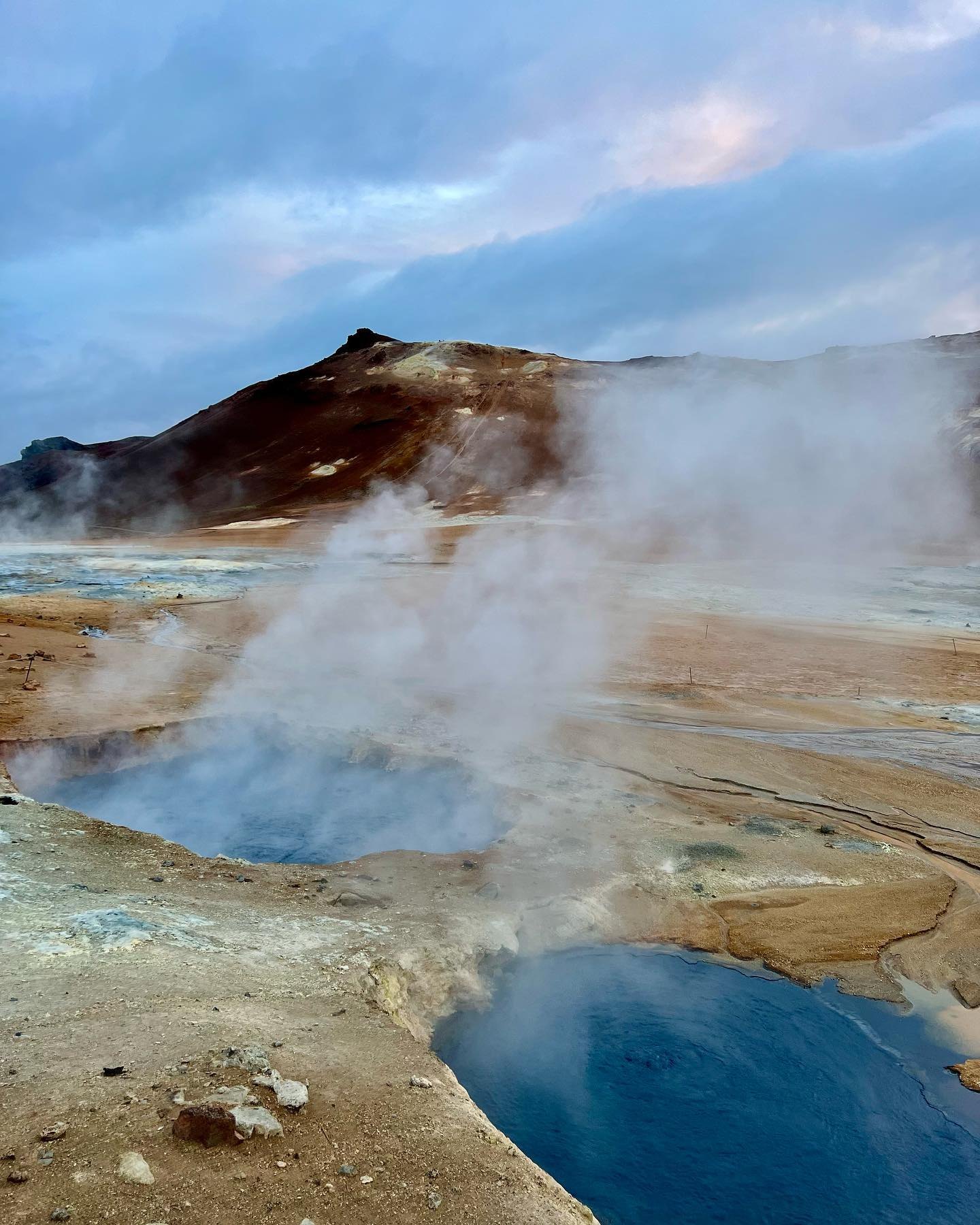 Steam drifting over the ochre ground of Námafjall Hverir and a milky blue pool, below a rust-red hill