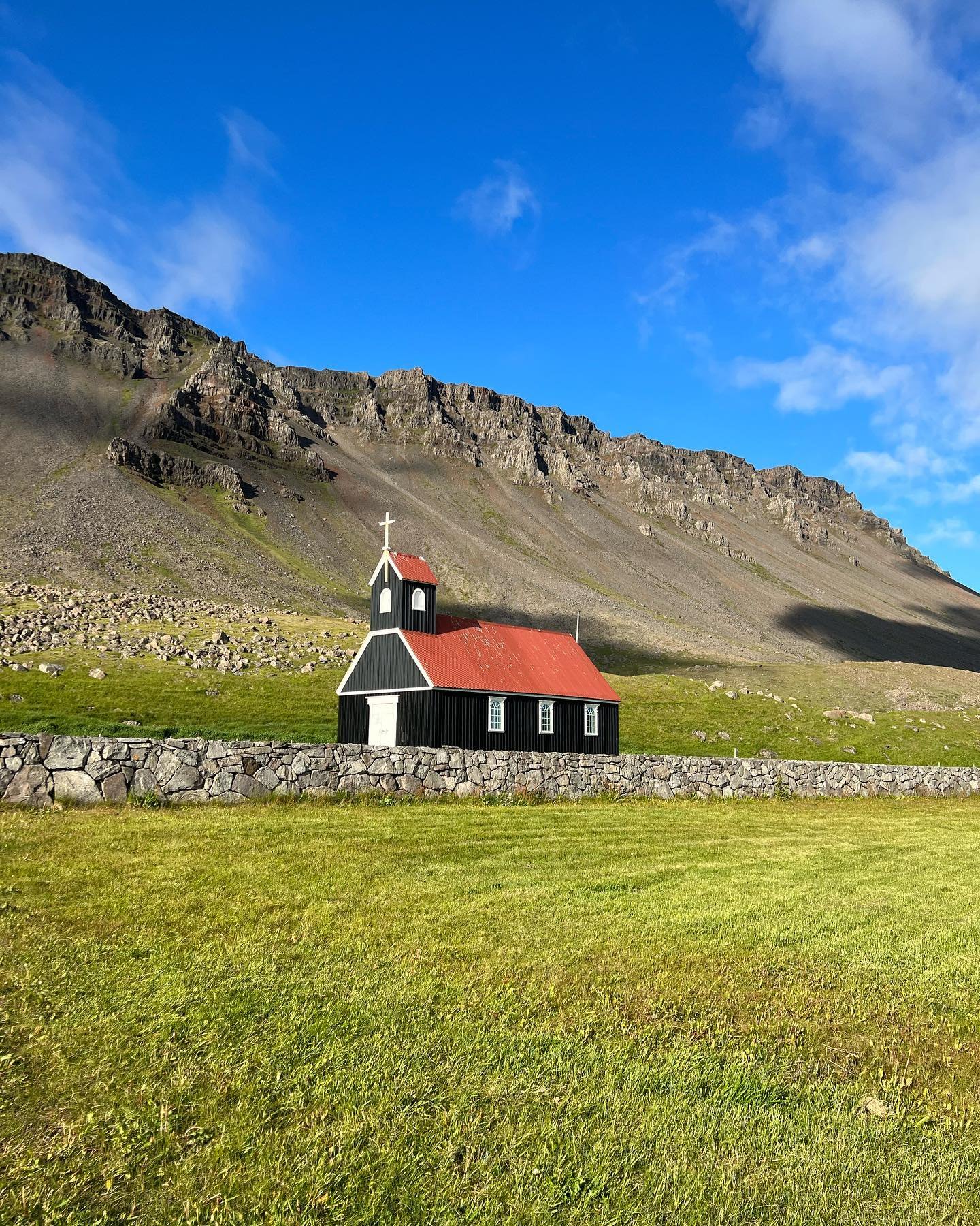 The small black church at Rauðasandur with its red roof, behind a stone wall below sunlit cliffs