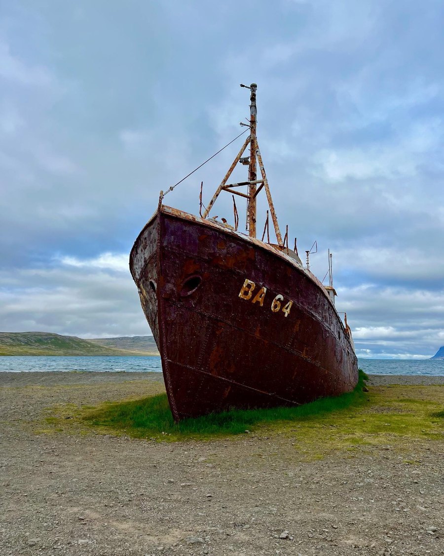 A rusting steel ship, BA 64 on its bow, beached on gravel and grass by the water